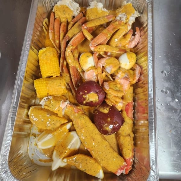 A seafood boil in a foil pan with crab legs, shrimp, corn, potatoes, and eggs, all covered in sauce.